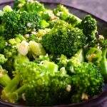 A closeup of stir fried broccoli on a dark blue platter, topped with sesame seeds and sitting on a dark blue marbled background.