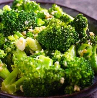 A closeup of stir fried broccoli on a dark blue platter, topped with sesame seeds and sitting on a dark blue marbled background.