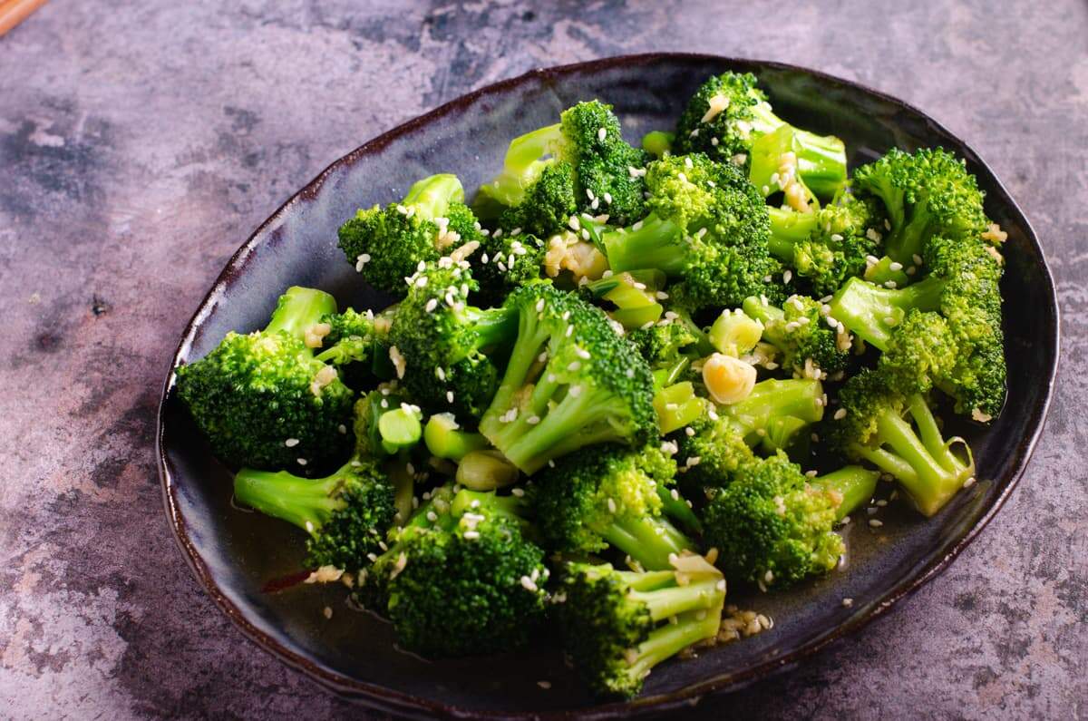 Stir fried broccoli on a dark blue platter, topped with sesame seeds and sitting on a dark blue marbled background.