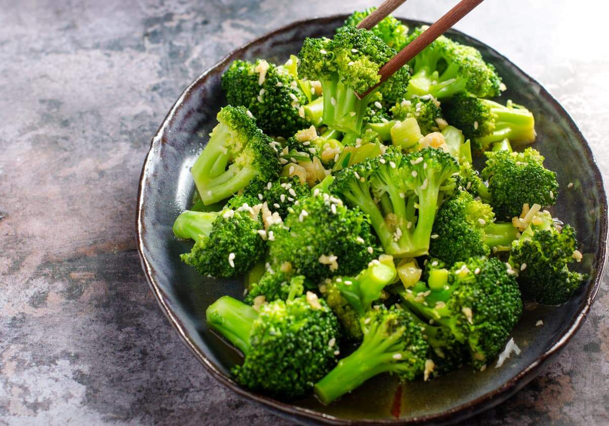 Stir fried broccoli on a dark blue platter, topped with sesame seeds and sitting on a dark blue marbled background and a hand using chopsticks to pick up a piece of the broccoli.