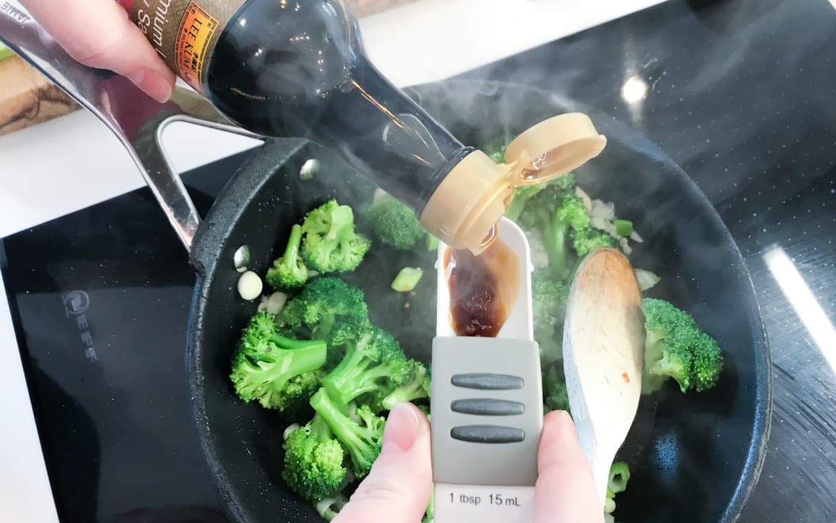 Adding a spoon of soy sauce to broccoli in a hot pan to stir fry.
