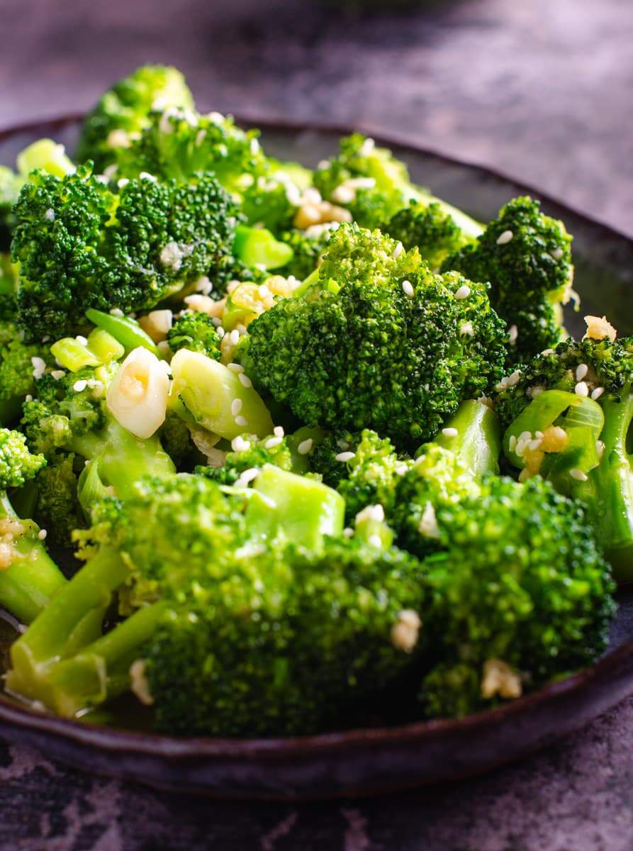 A closeup of stir fried broccoli on a dark blue platter, topped with sesame seeds and sitting on a dark blue marbled background.
