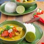 Two green bowls on matching plates filled with vegetable and coconut curry served with rice, limes and scattered green onions and fresh chillies.
