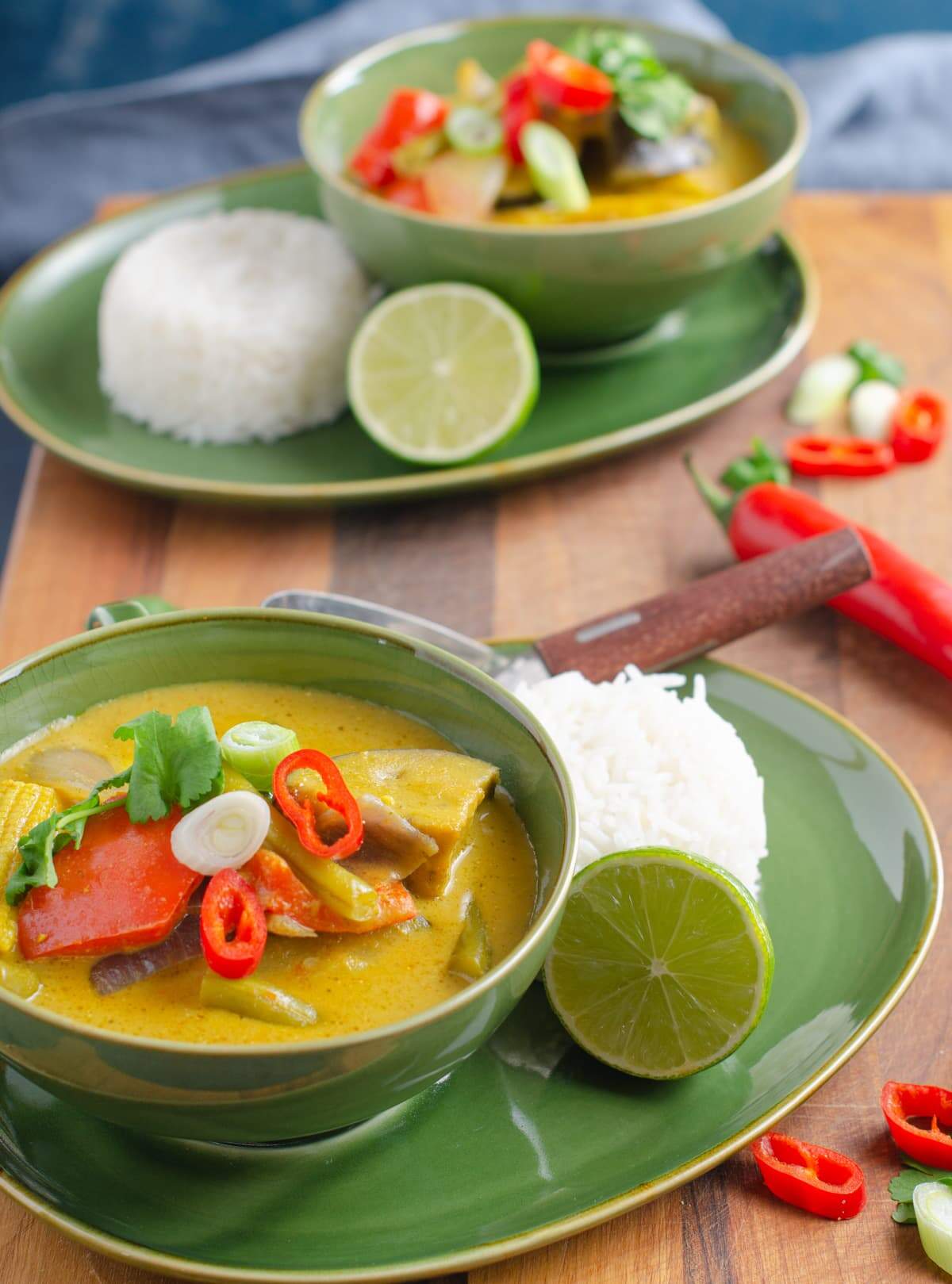Two green bowls on matching plates filled with vegetable and coconut curry served with rice, limes and scattered green onions and fresh chillies.