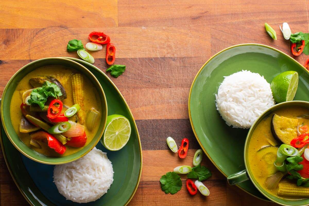 A flat lay image of 2 green bowls on matching plates filled with vibrant vegetable and coconut curry served with portions of rice and some fresh coriander and chillies scattered around.