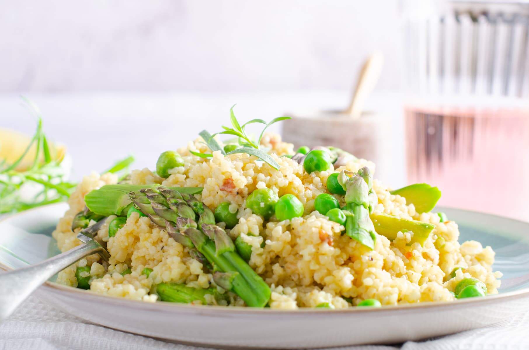 A frontal view of a plate of bulgar wheat risotto, a white waffled napkin, lemon and tarragon to the back and a glass of rose to the right.