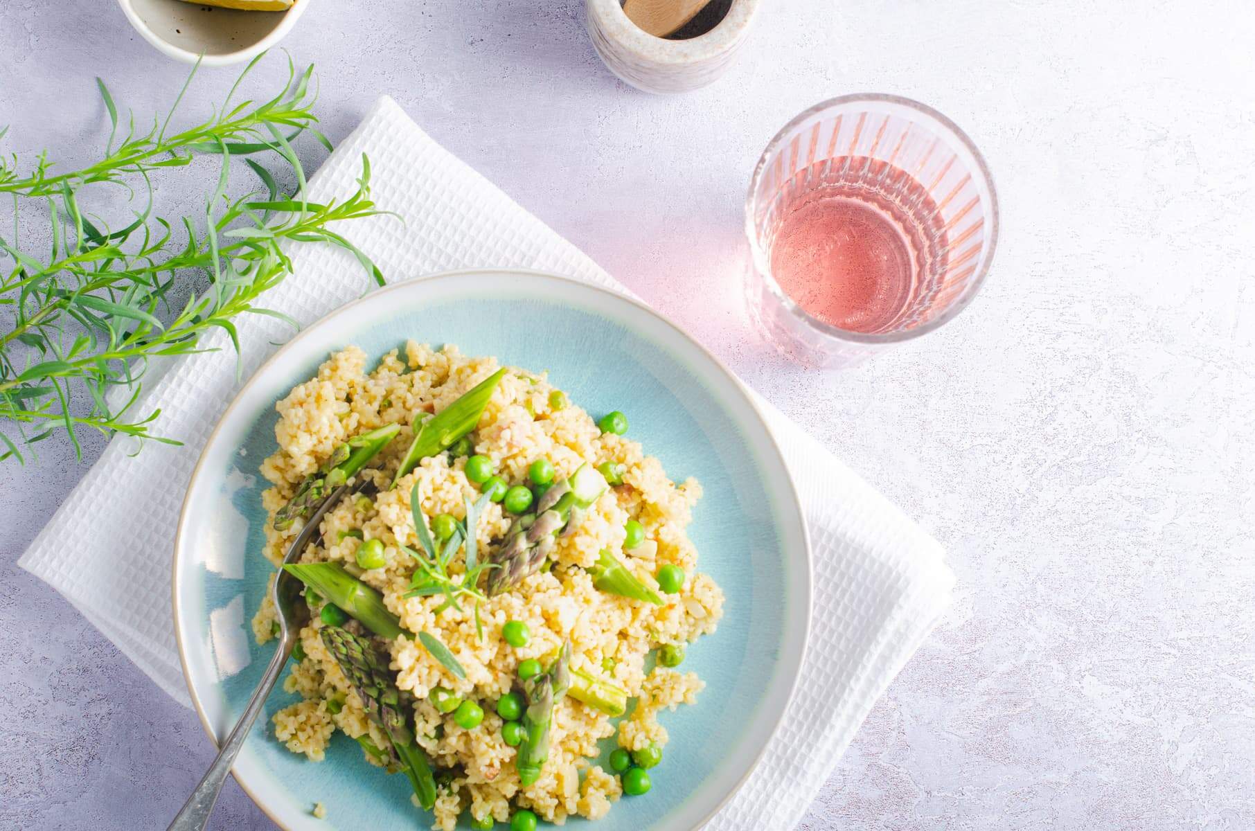 A top down view of a spring bulgar wheat risotto with peas, asparagus and topped with tarragon, a glass of rose to the right and tarragon and lemon to the left.
