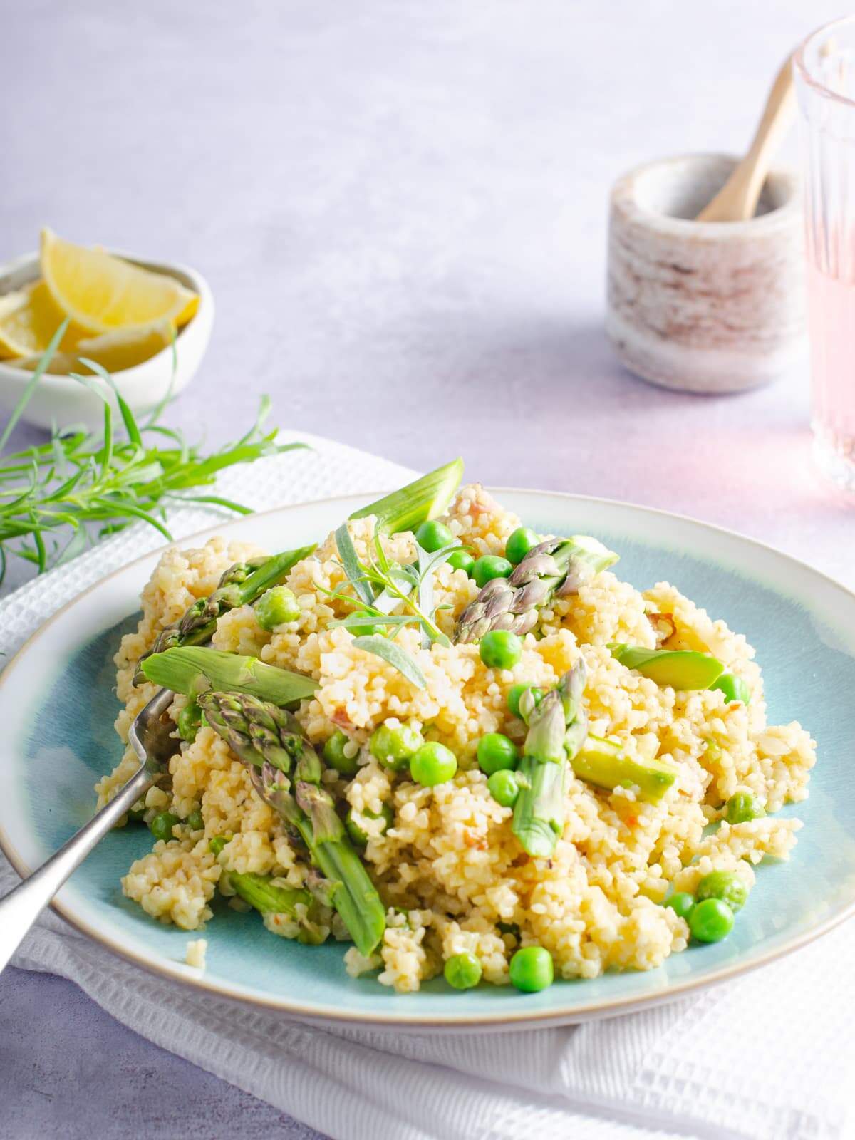 A blue mottled plate with bulgar wheat risotto with asparagus, peas and tarragon sitting on a white napkin served with a glass of rose wine.