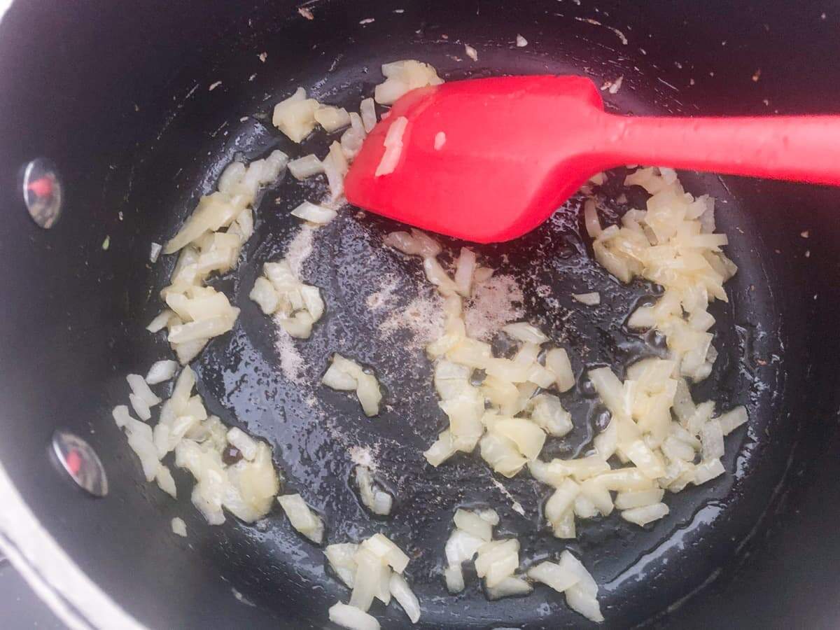 A black fry pan cooking diced onions and a red spatula stirring them.