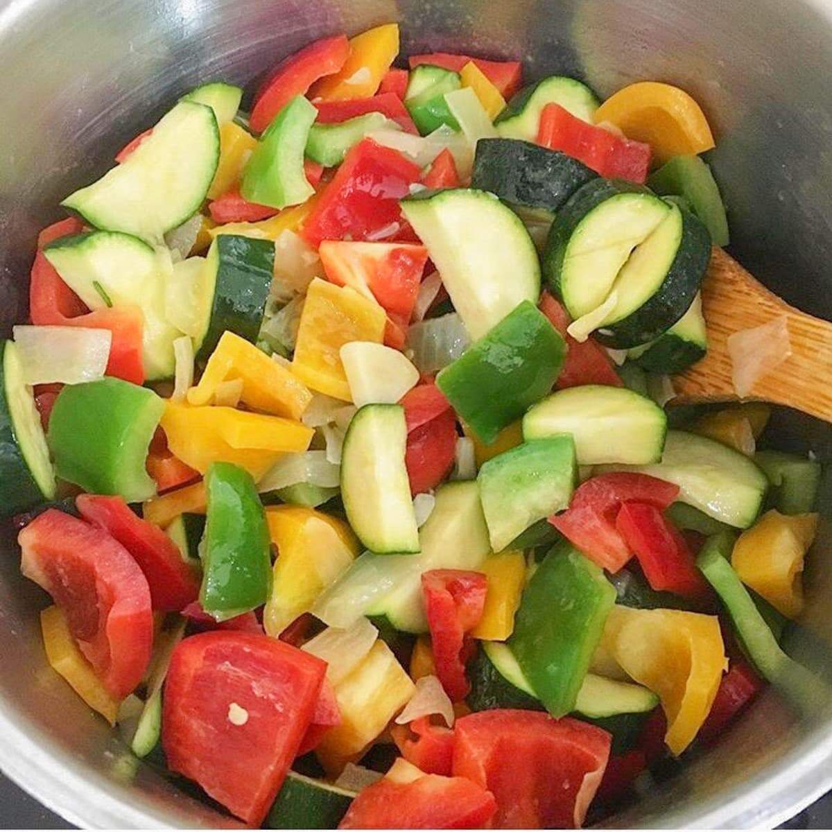 Chunky chopped courgettes (zucchini), red, yellow and green peppers with sautéed onions ready for soup making in a large pan.
