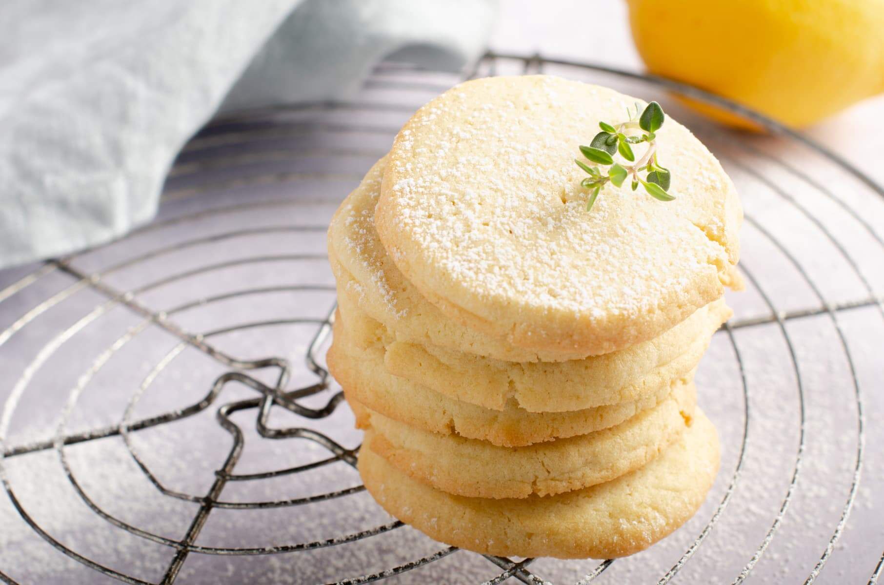 A tower of simple lemon butter cookies sat on top of a wire cooling rack, topped with powdered sugar and a snip of lemon thyme with a lemon and pale blue napkin to the back.