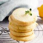 A tower of simple lemon butter cookies sat on top of a wire cooling rack, topped with powdered sugar and a snip of lemon thyme with a lemon to the back.