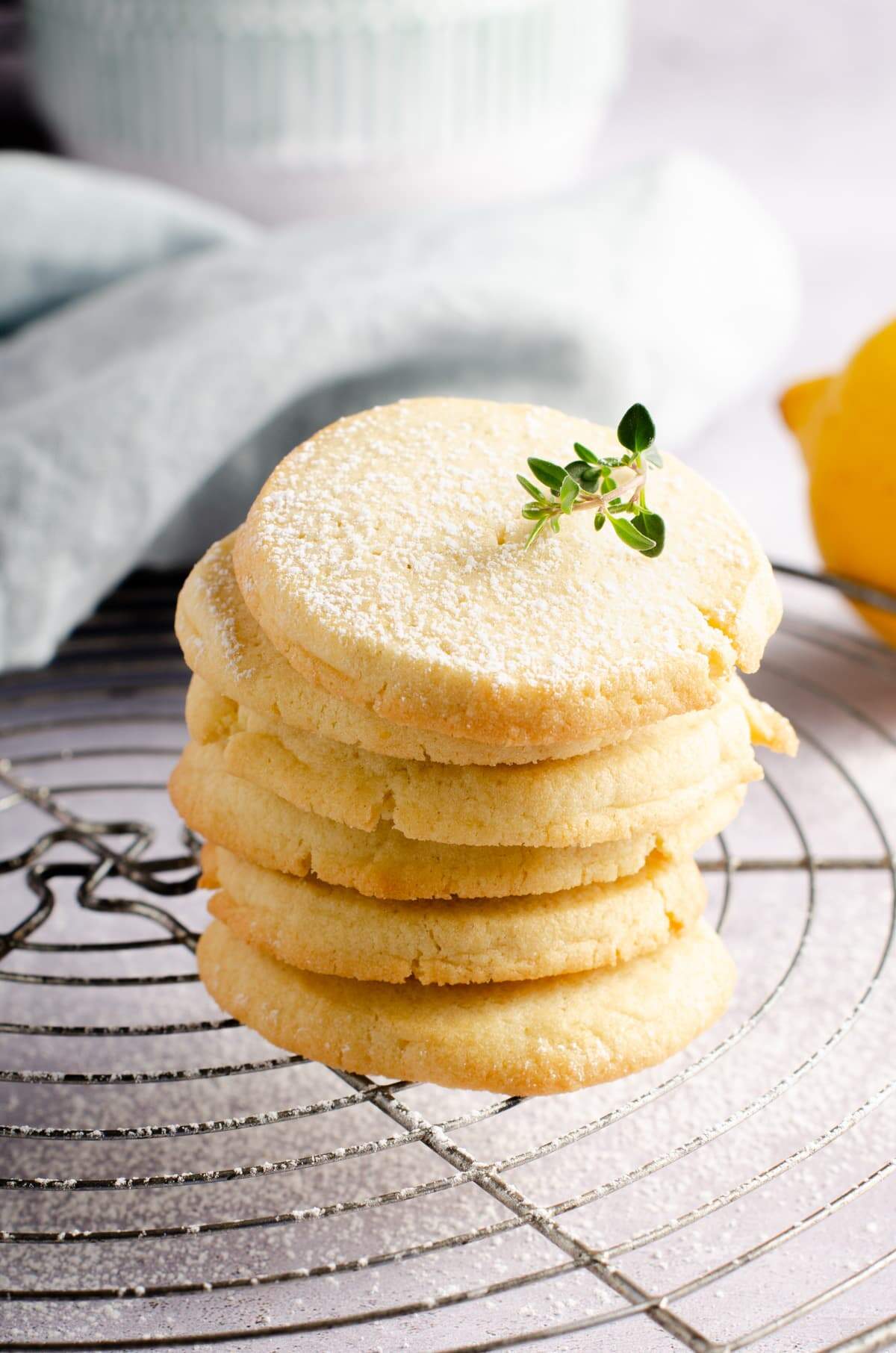 A tower of simple lemon butter cookies sat on top of a wire cooling rack, topped with powdered sugar and a snip of lemon thyme with a lemon to the back.