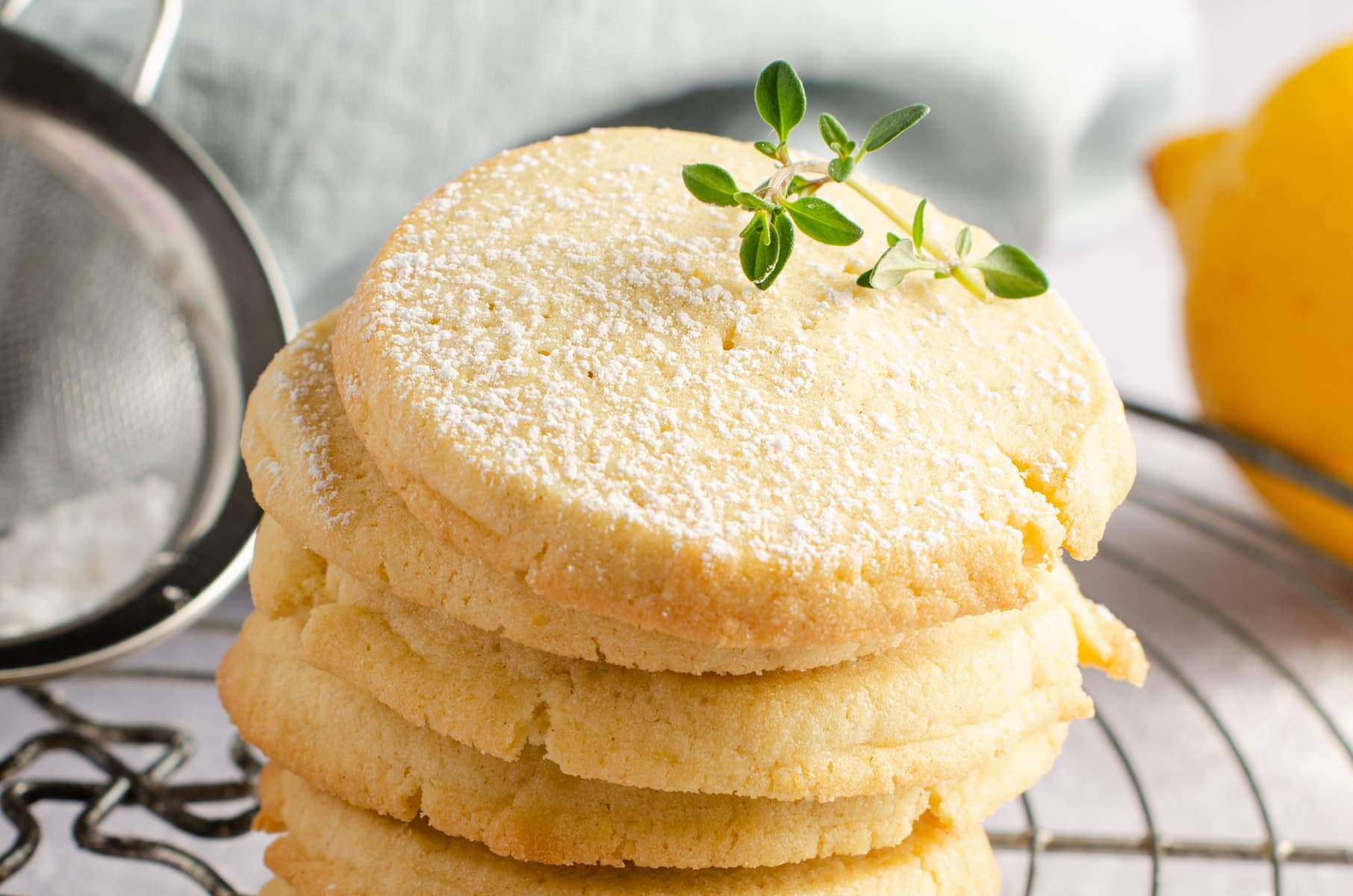 Delicious and simple lemon butter cookies topped with icing sugar and a sprig of lemon thyme on a wire cooling rack, some lemons to the back and the icing sugar shaker to the side