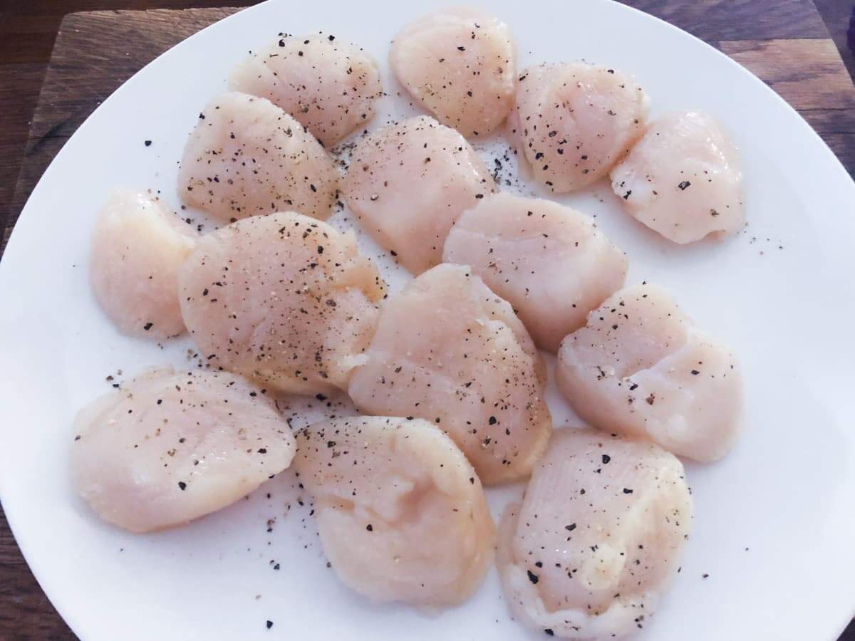 Scallops seasoned with salt and black pepper on a white plate before being cooked.