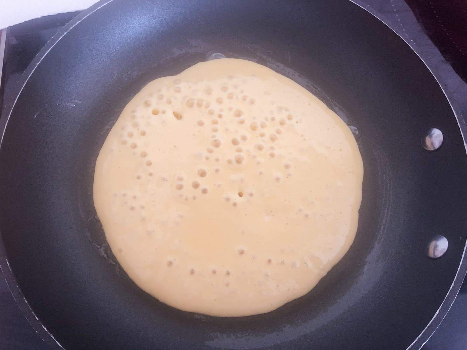 A pancake cooking in a black frying pan ready to be flipped with bubbles showing on the top.