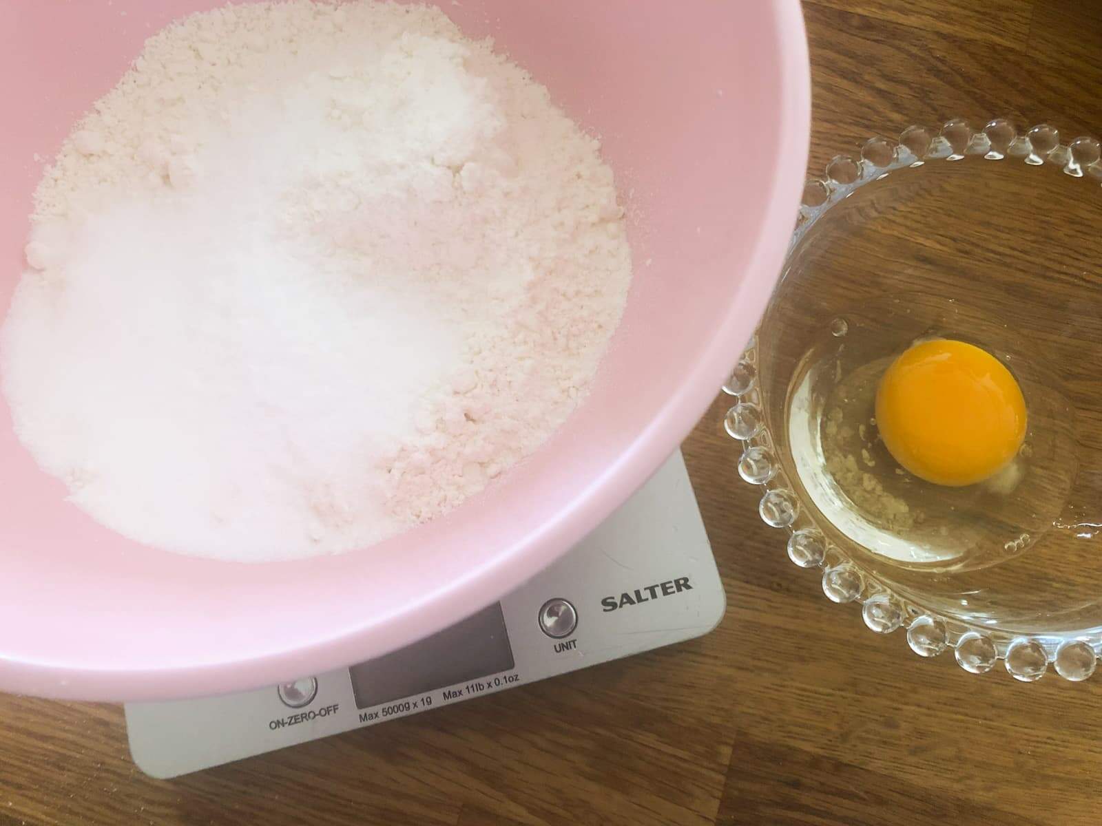 A set of scales with a pink bowl containing flour and sugar weighed out and a smaller glass bowl with an egg ready to be mixed together.