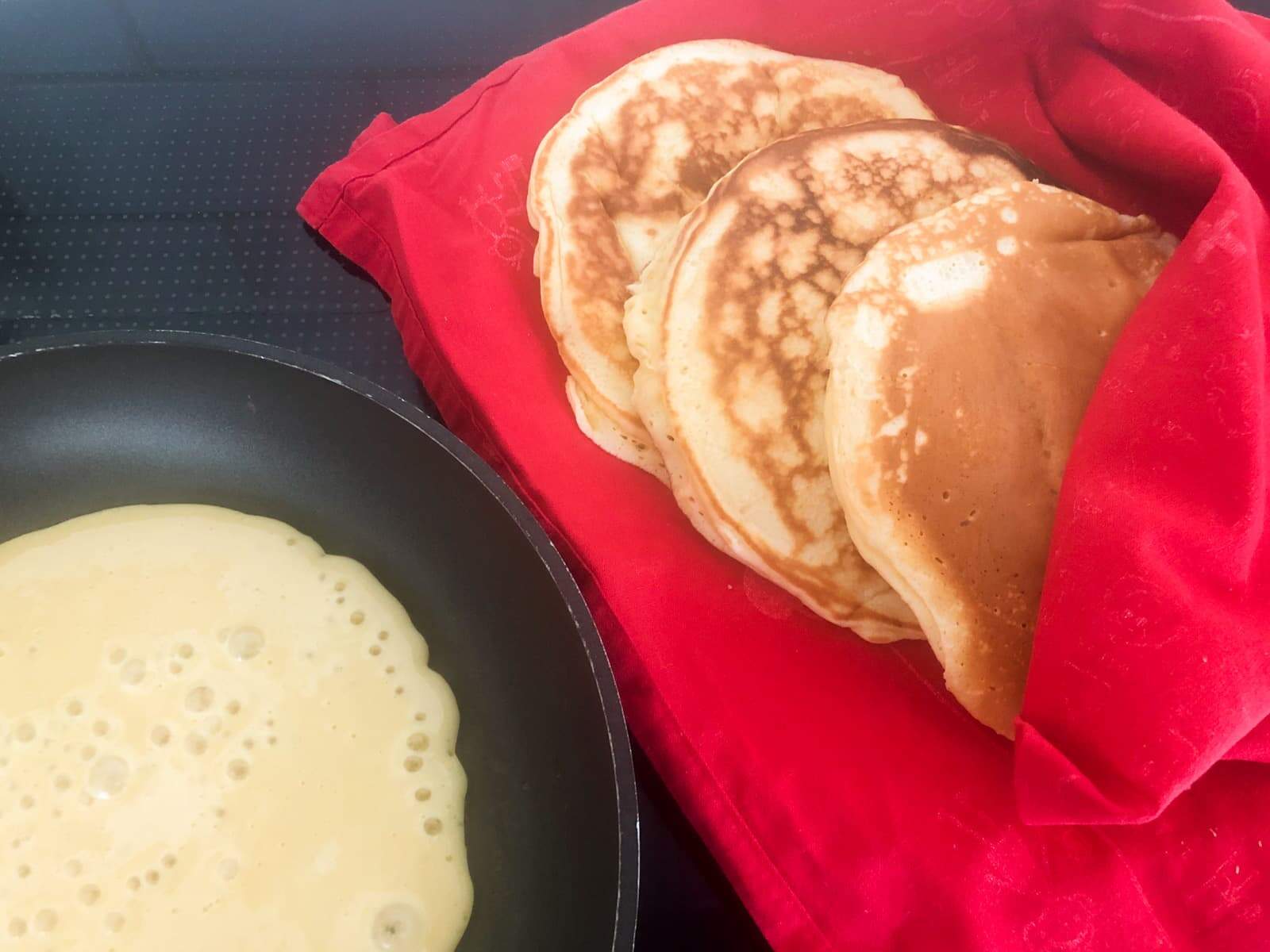 A black frying pan cooking pancakes and a red tea towel to the right holding the already cooked pancakes and keeping them warm.