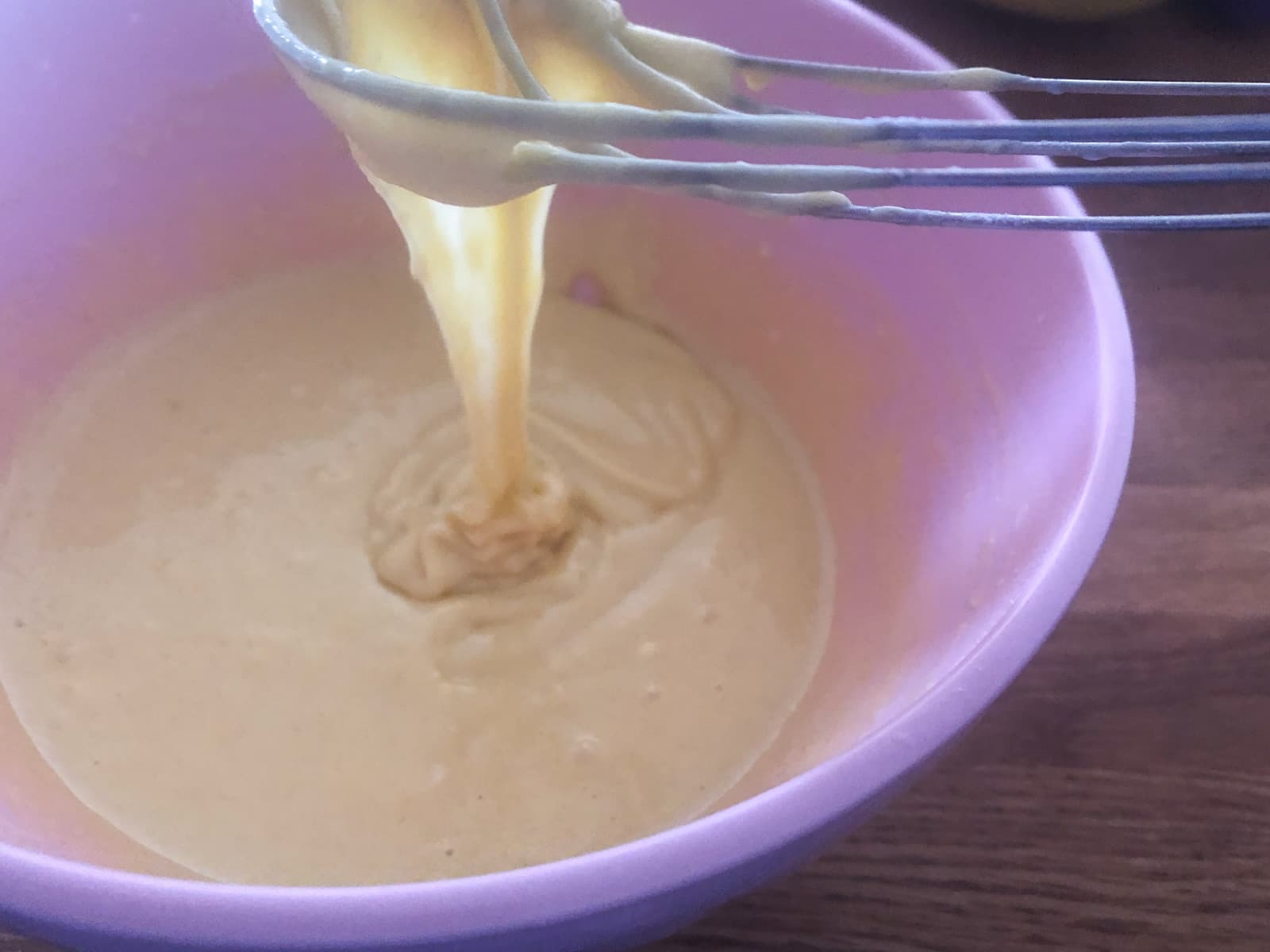 A pink bowl with a pancake batter and a whisk with the batter dropping from it to show the consistency of the batter.