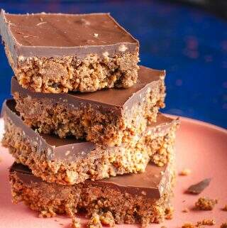 A stack of 4 chocolate tiffin squares on a salmon pink plate and lots of crumbs on a dark blue back drop.