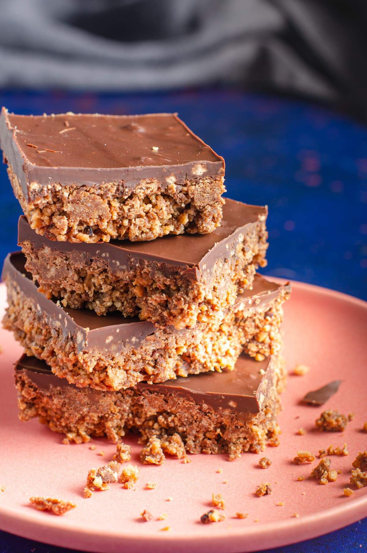 A stack of 4 chocolate tiffin squares on a salmon pink plate and lots of crumbs on a dark blue back drop.