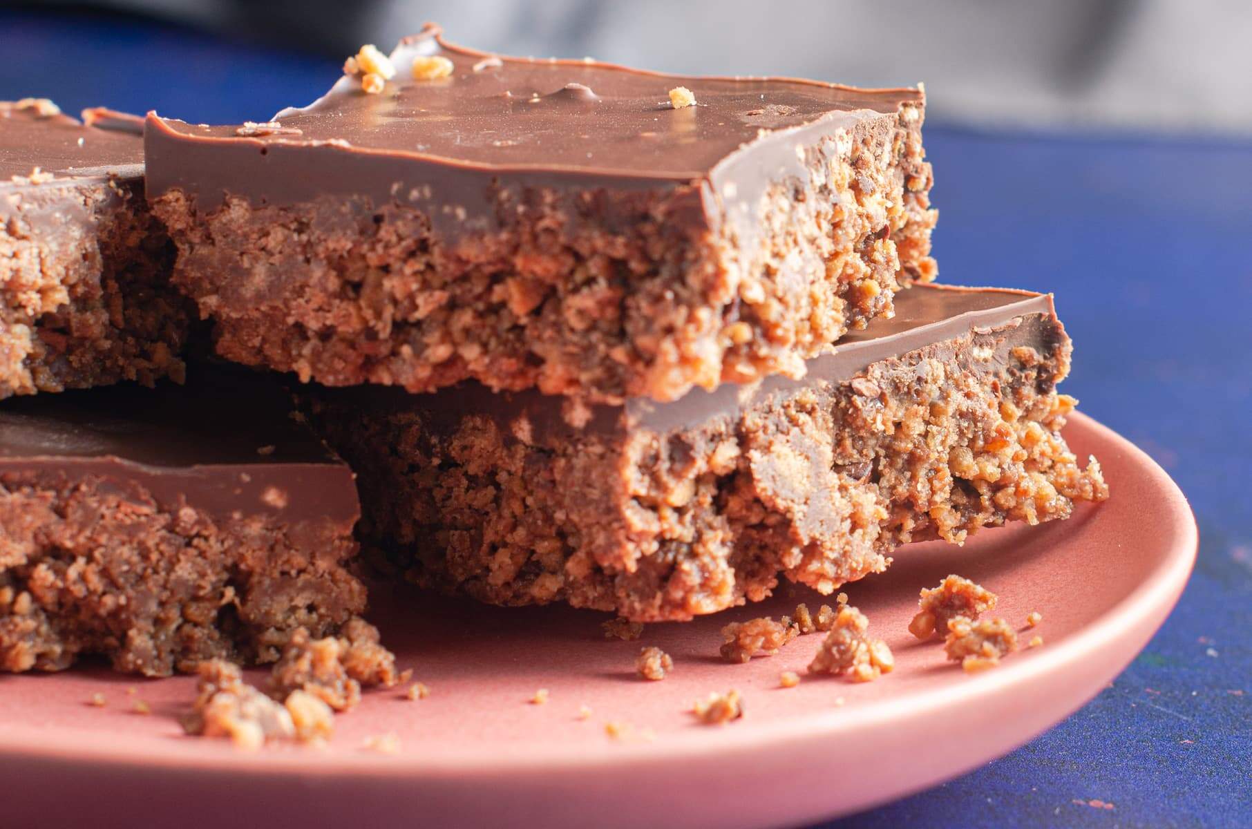 4 squares of crumbly no bake chocolate tiffin digestive traybake sat on a salmon pink coloured plate, crumbs scattered and a dark blue backgroiund