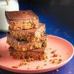 A stack of 4 chocolate no bake digestive tiffin squares on a salmon pink plate with an old school milk bottle and straw to the back left and lots of crumbs on a dark blue back drop.
