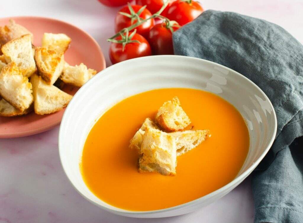 A bowl of creamy vegan tomato soup topped with crunchy sourdough croutons and a bunch of red vine tomatoes in the back.