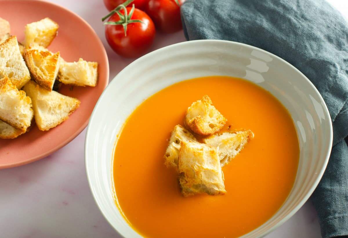 A bowl of creamy vegan tomato soup topped with crunchy sourdough croutons and a bunch of red vine tomatoes in the back.