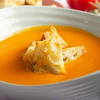 A close up of a bowl of creamy vegan tomato soup topped with crunchy sourdough croutons and a bunch of red vine tomatoes in the back.