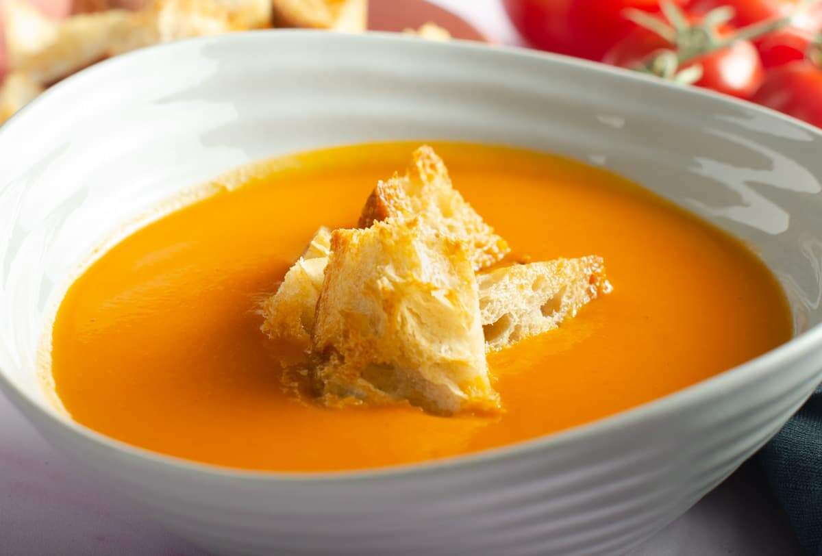 A close up of a bowl of creamy vegan tomato soup topped with crunchy sourdough croutons and a bunch of red vine tomatoes in the back.