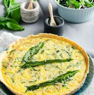 A wild garlic, cheese and onion quiche on a blue plate with fresh wild garlic leaves, a green salad and salt and pepper pots to the back.