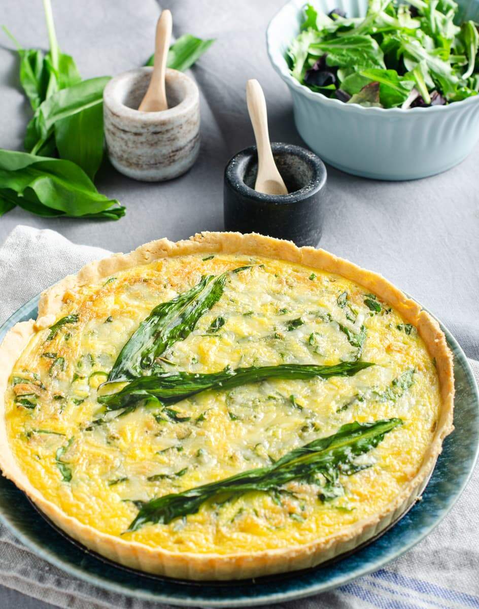 A wild garlic, cheese and onion quiche on a blue plate with fresh wild garlic leaves, a green salad and salt and pepper pots to the back.