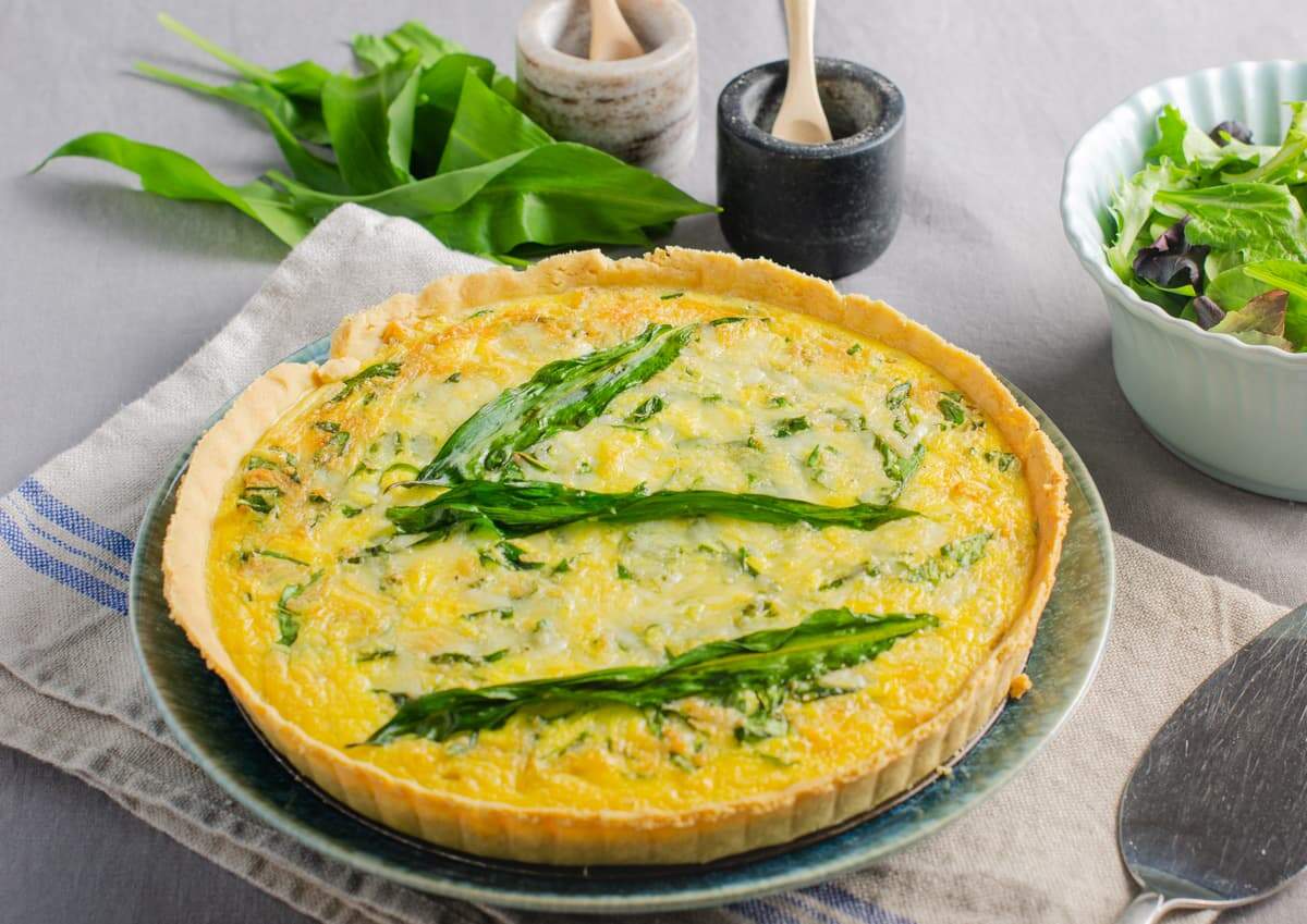 A table setting with a wild garlic, cheese and onion quiche on a blue plate, a serving slice to the side, some green salad and sat on a grey tablecloth with a lined striped napkin underneat.