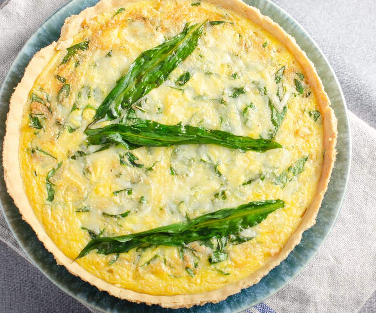 A top down view of a wild garlic, cheese and onion quiche on a blue plate and say on a grey tablecloth with a linen napkin.