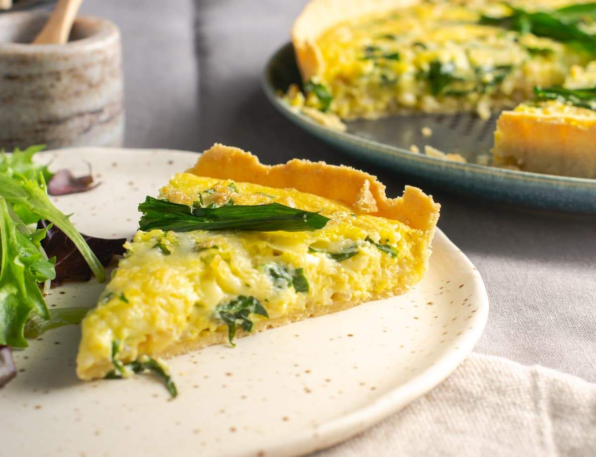 A serving of wild garlic, cheese and onion quiche with a side of a green salad on a speckled plate and the rest of the quiche to the back with salt and pepper pots to the back.