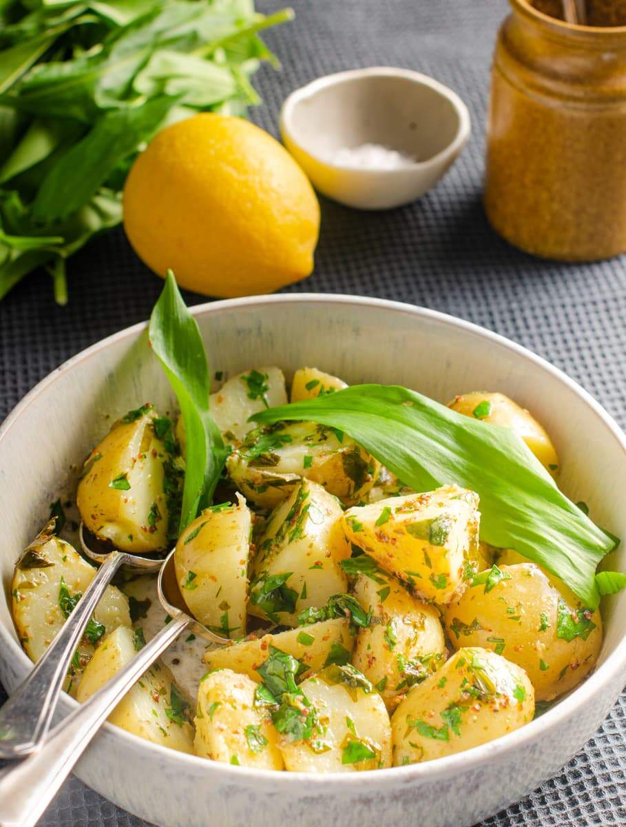 A mottled ceramic bowl filled with warm wild garlic potato salad, with a lemon to the back, fresh picked wild garlic and a mustard pot on a dark blue tablecloth
