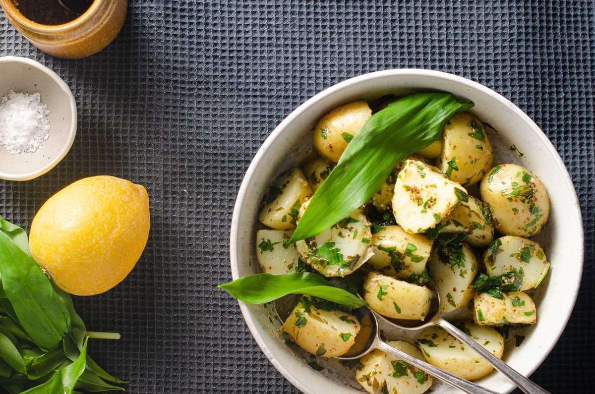 An image from above of a bowl of warm potato salad with wild garlic, lemon and mustard on a blue waffled tablecloth.