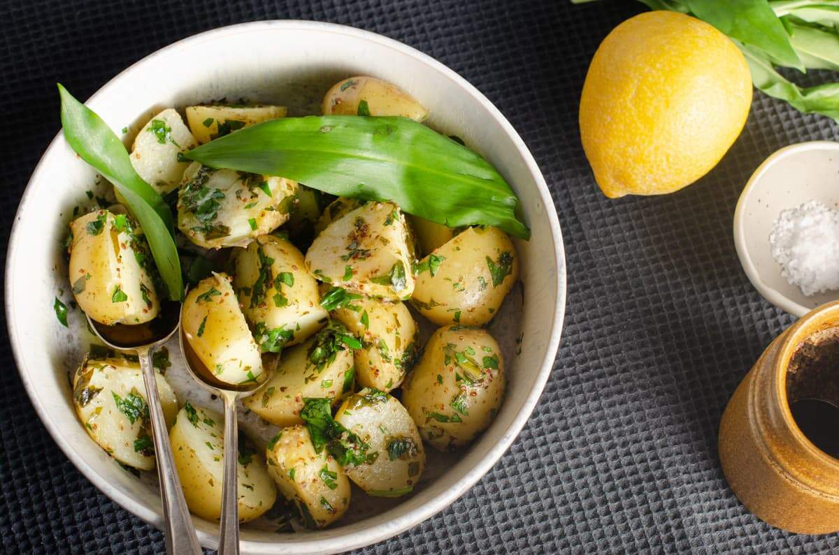 An inviting bowl of warm wild garlic potato salad with fresh wild garlic leaves tossed in, a lemon, mustard and fresh picked leaves just to the side.