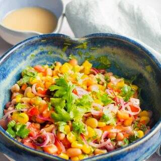 A heart shaped blue mottled ceramic bowl filled with a colourful mixed bean salad with a pouring jug of vinaigrette behind.