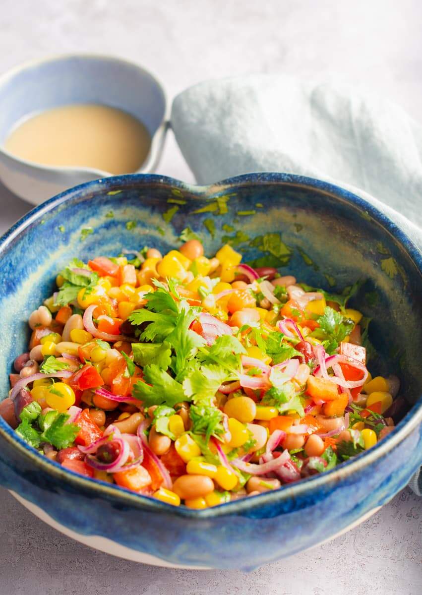 A heart shaped blue mottled ceramic bowl filled with a colourful mixed bean salad with a pouring jug of vinaigrette behind.