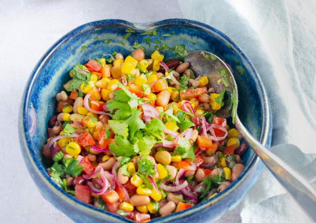 A blue ceramic heart shaped bowl on a grey surface filled with a vibrant colourful mixed bean salad.