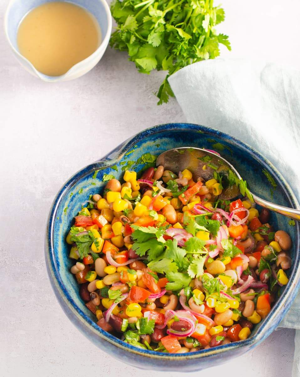 A heart shaped blue mottled ceramic bowl filled with a colourful mixed bean salad with a pouring jug of vinaigrette behind with a handful of fresh coriander.