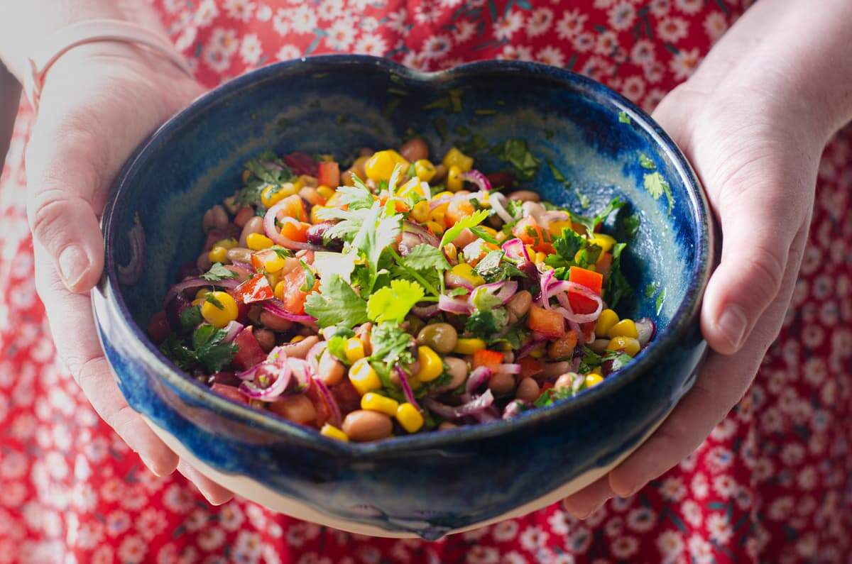 A young girl holding a heart shaped bowl filled with a mixed bean salad and wearing a red and white flowered dress.