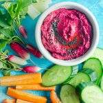 A bright blue table setting with a tub of beetroot hummus and lots of fresh crunchy vegetables surrounding including sliced cucumber, peppers, carrots, celery and radish.