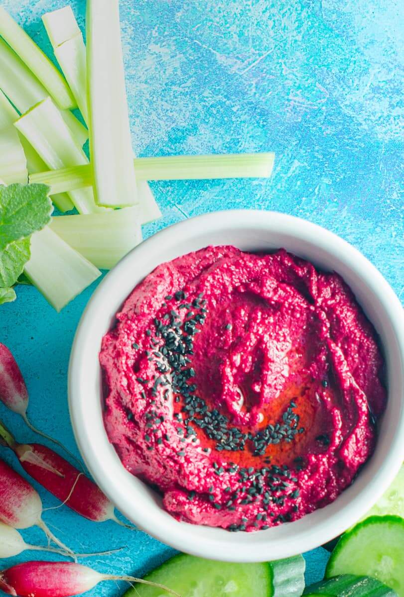 A dish of beetroot hummus with black nigella seeds on top in a white tub and lots of fresh vegetables surrounding it on a bright blue surface.
