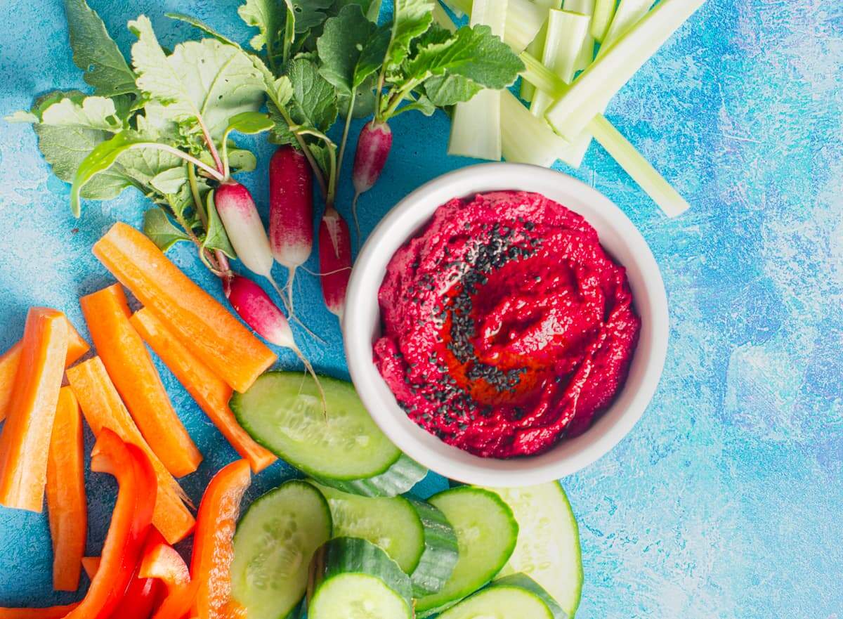 A flatlay of beetroot hummus with radish, carrot sticks, sliced cucumber and red peppers surrounding on a bright blue surface.