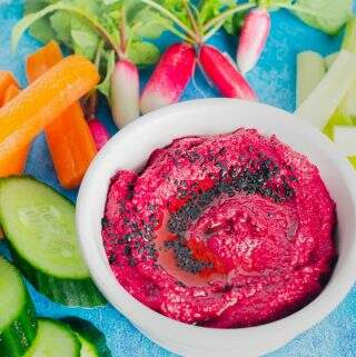 A white tub containing beetroot hummus topped with nigella seeds and fresh raw vegetables near it.