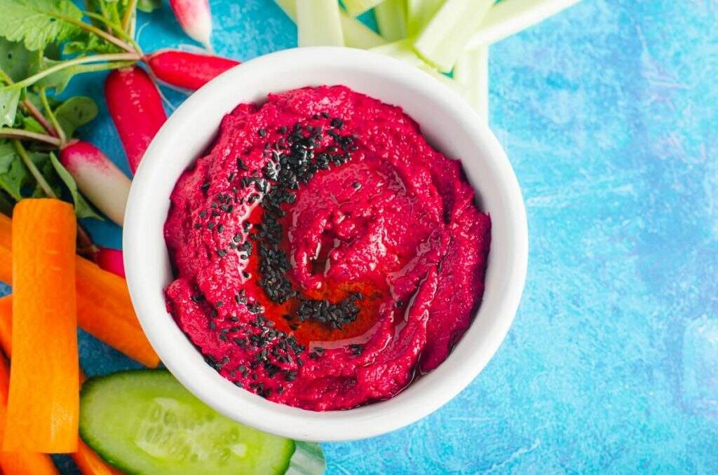 A top down view of a bowl of beetroot hummus with fresh vegetable for dipping.