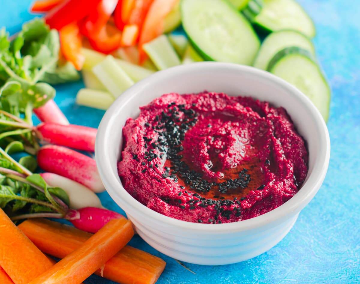 Fresh vibrant beetroot hummus topped with nigella seeds and surrounded by fresh vegetable to dip.